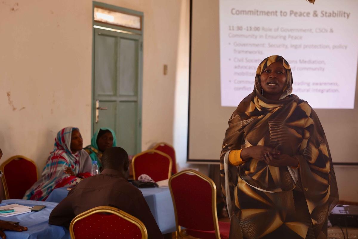 A woman addressing a group of people seated at tables. In the background, there is a PowerPoint presentation. that the woman is presenting.