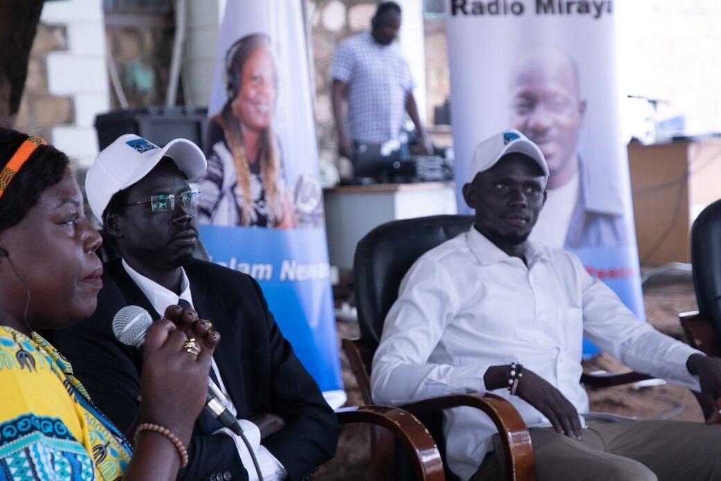 Three people seated indoors, one holding a microphone, with banners and a person standing near audio equipment in the background.