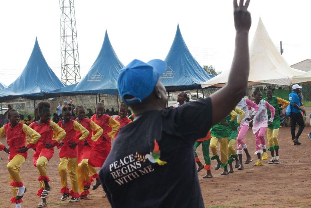 Outdoor event with a person wearing a shirt that reads “Peace Begins With Me” raising a hand, while groups of performers in colorful outfits dance in formation near blue tents.