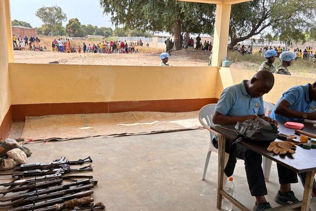 Deux hommes sont assis à une table, à côté d'une rangée d'armes à feu posées sur le sol.